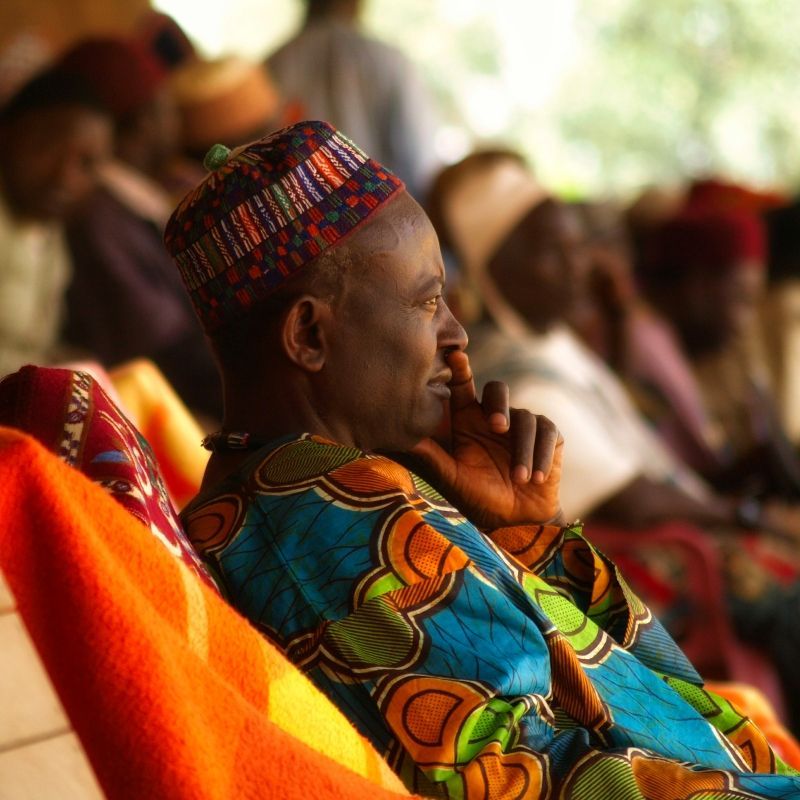 Man in colorful patterned clothing and cap, seated outdoors, deep in thought.