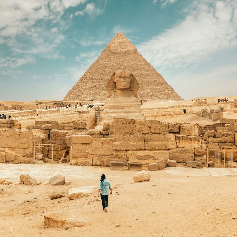 A woman is walking in front of a pyramid and a sphinx statue.