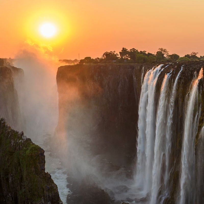Experience the awe of Victoria Falls with Inspiration Africa. This stunning image captures the mighty waterfall at sunset, with golden light reflecting off the cascading waters and mist rising into the sky. Inspiration Africa offers expertly curated trips to explore this natural wonder, known locally as Mosi-oa-Tunya, or