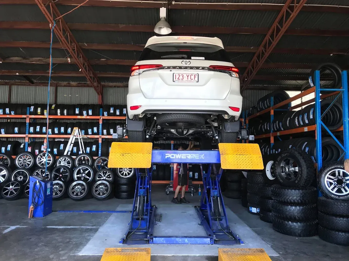 A white SUV elevated on a blue hydraulic lift inside a tire shop filled with rows of stacked tires.