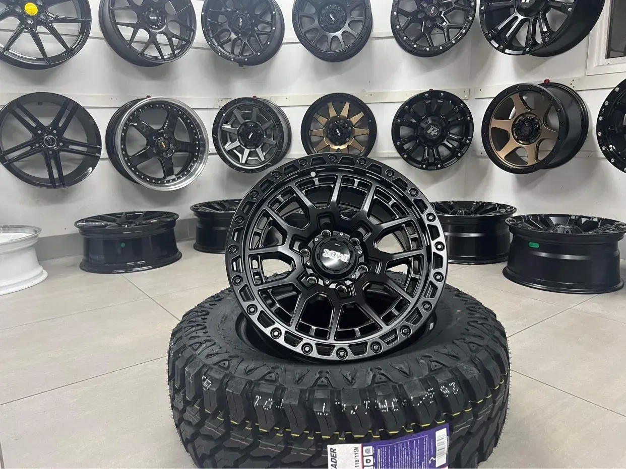 A large, black off-road tire mounted with a textured black wheel, displayed in front of a wall of various car wheel rims.