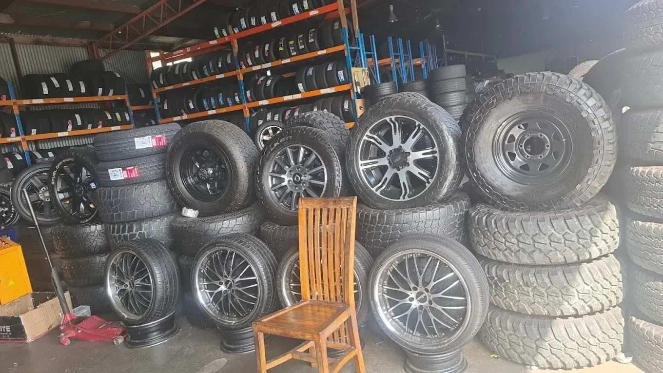 A wooden chair sits in front of a cluttered tire shop filled with stacks of various automotive tires and custom rims.