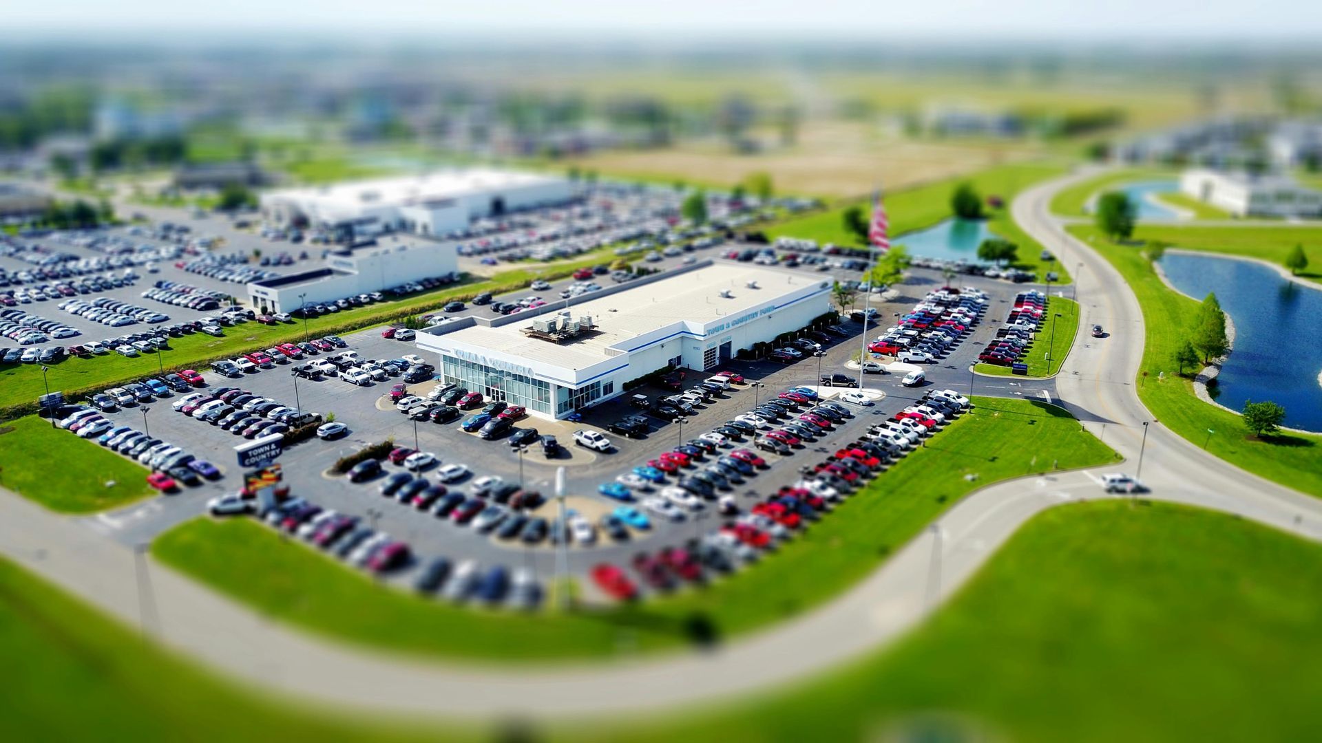 Car dealership with rows of vehicles, white building, green grass, and a pond.