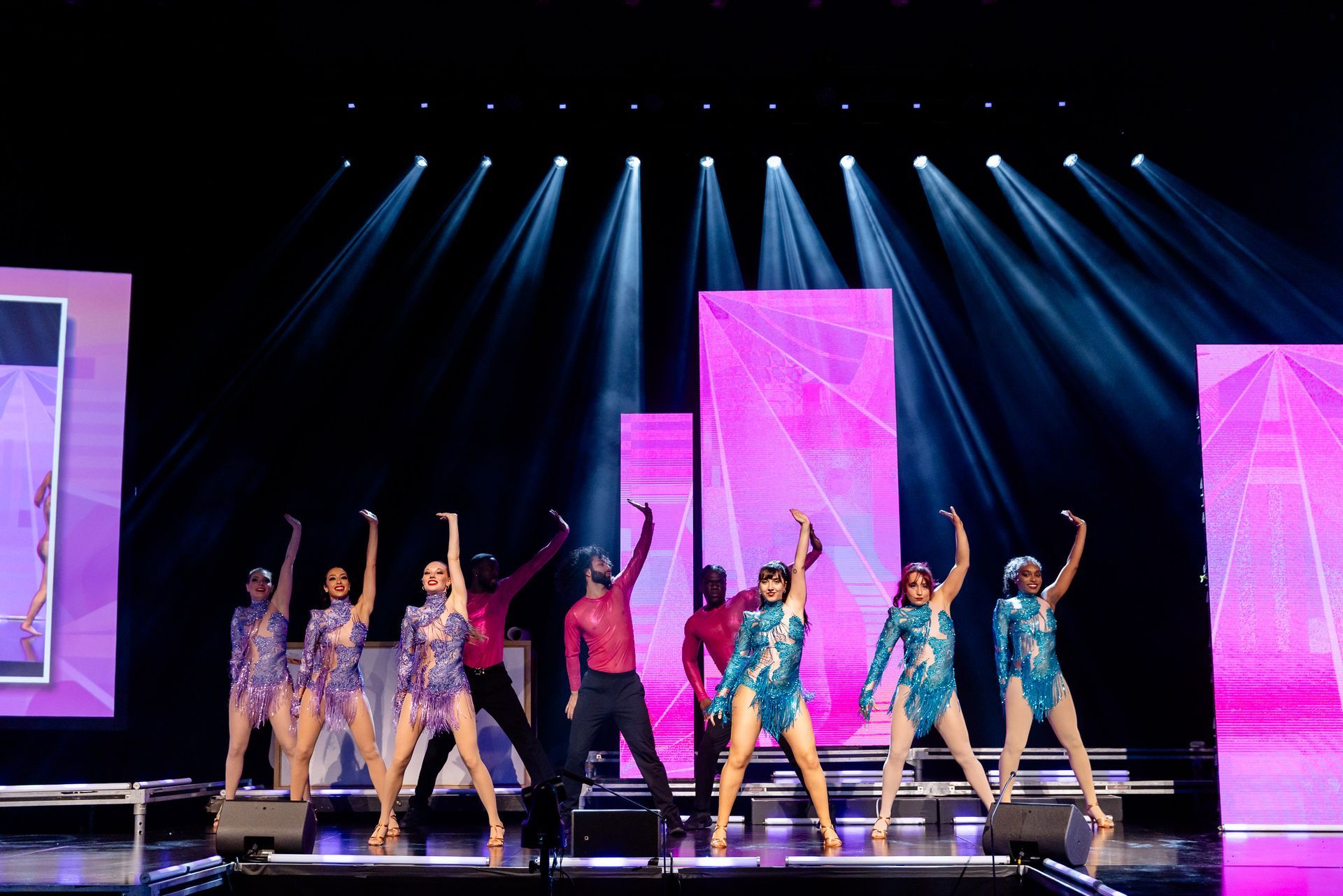 Dancers on stage with arms raised under spotlights, dressed in colorful costumes, with pink backdrop.