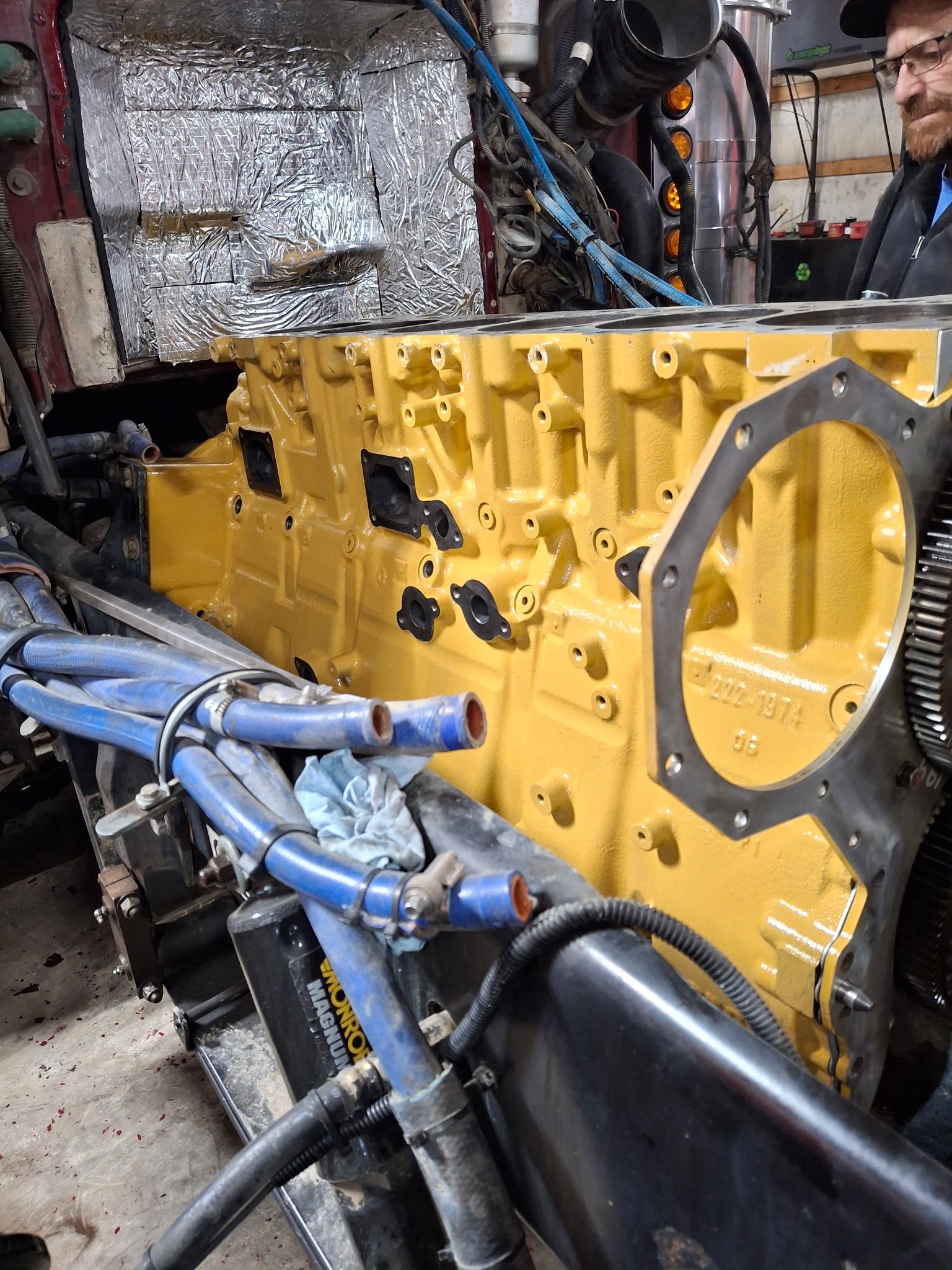 A worker inspecting a large, yellow engine block inside a repair shop, with blue hoses visible in the foreground.