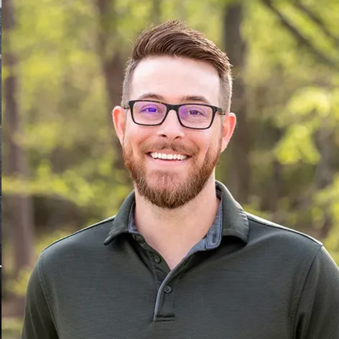 Man with glasses and beard smiles outdoors, wearing a green polo shirt.