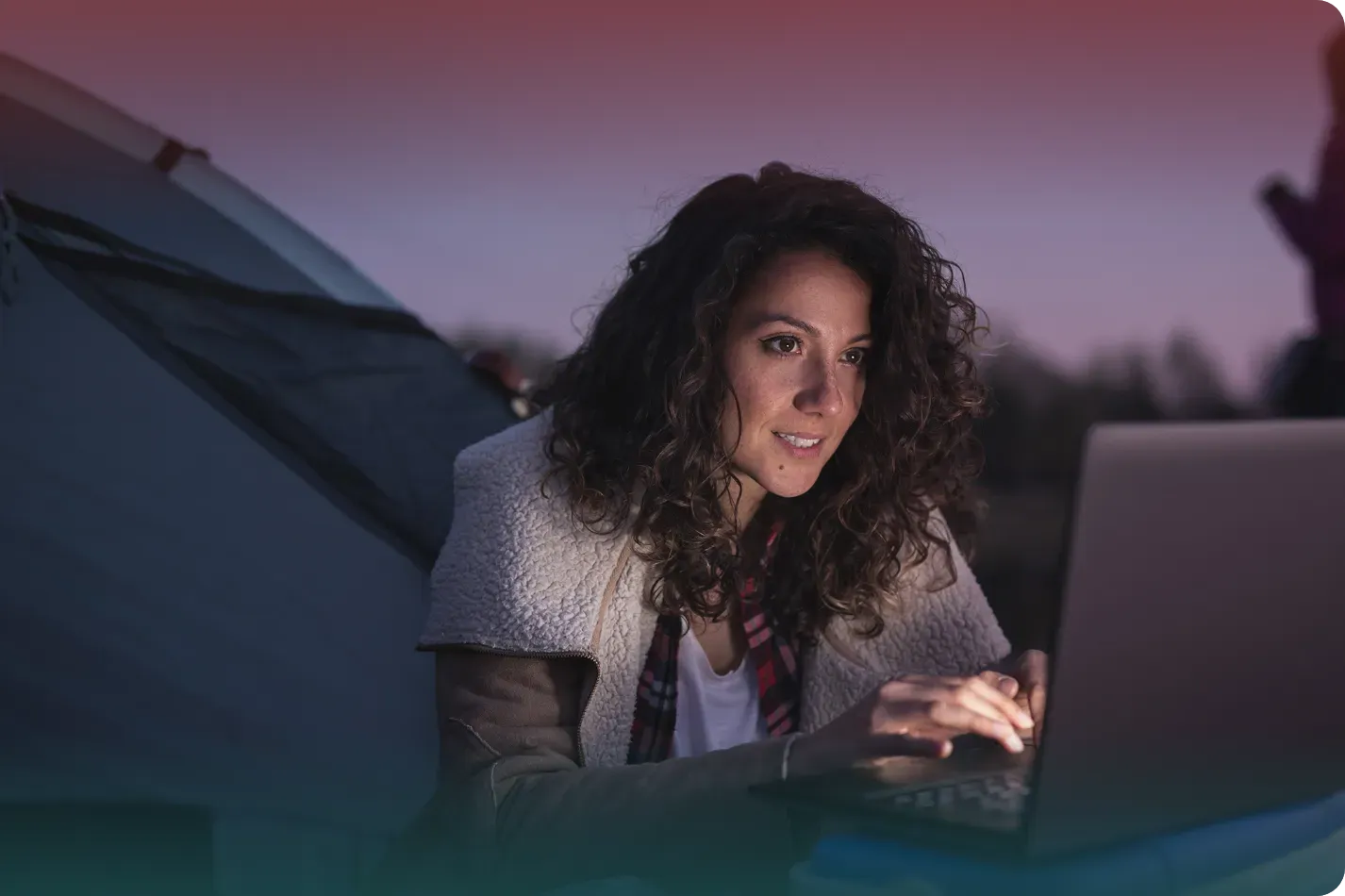 Woman with curly hair, lit by laptop, working outside a tent at dusk.