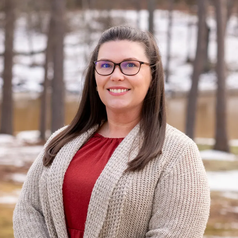 Woman in glasses and red top smiles, wearing a cream sweater, outdoors in front of trees.