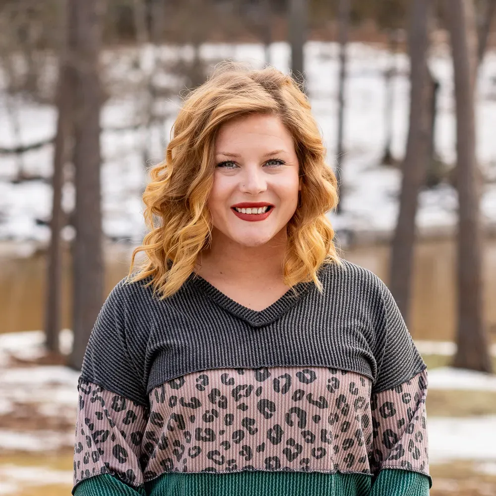 Woman with red hair, wearing a striped and leopard print top, smiling outdoors.