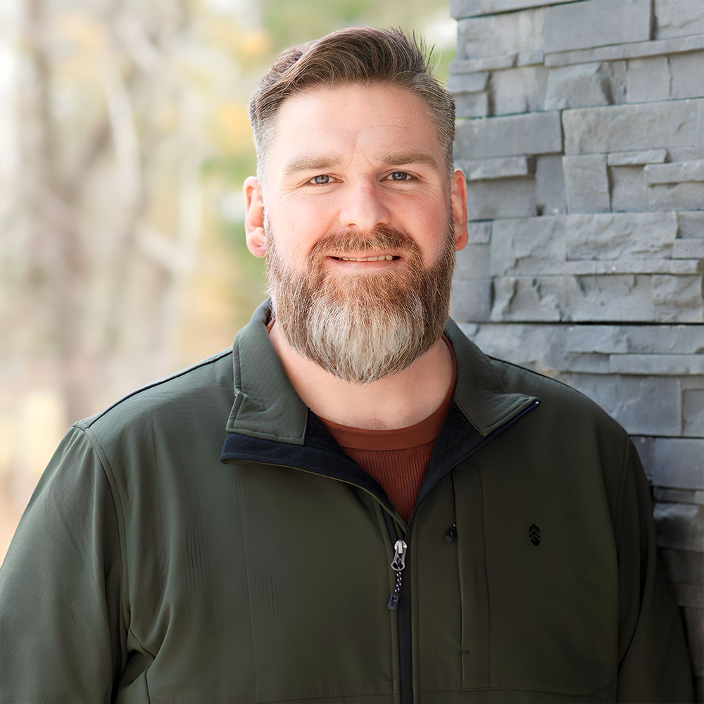 A man with a beard and a green jacket is standing in front of a brick wall.