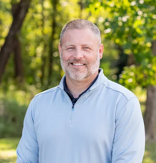 Man with graying beard smiles outdoors wearing a light blue quarter-zip in a green setting.