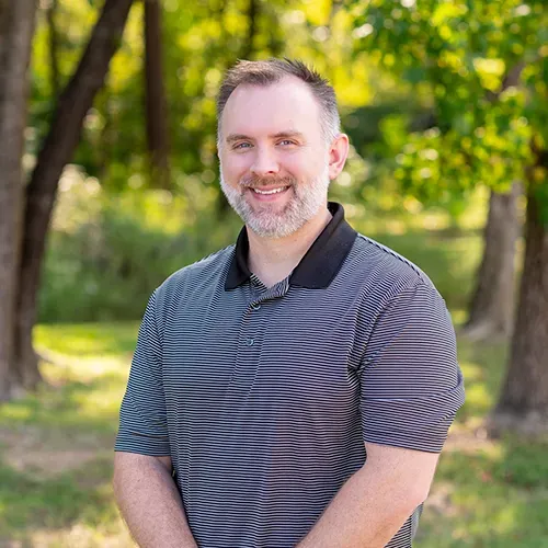 Man with graying beard, wearing a gray and black striped polo shirt, smiling in front of trees.
