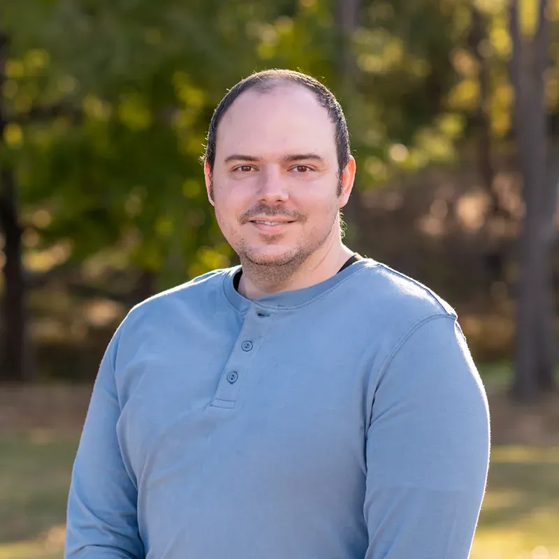 Man with light skin and a blue shirt smiles outdoors in front of trees.