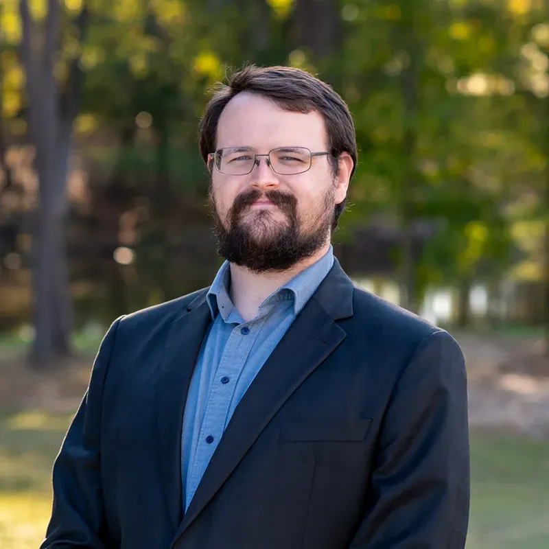 Man with glasses and beard in a blue shirt and black jacket, standing outside.