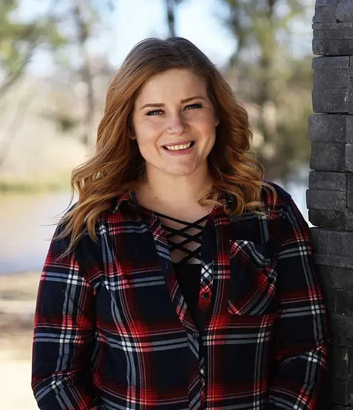 A woman in a plaid shirt is leaning against a brick wall and smiling.