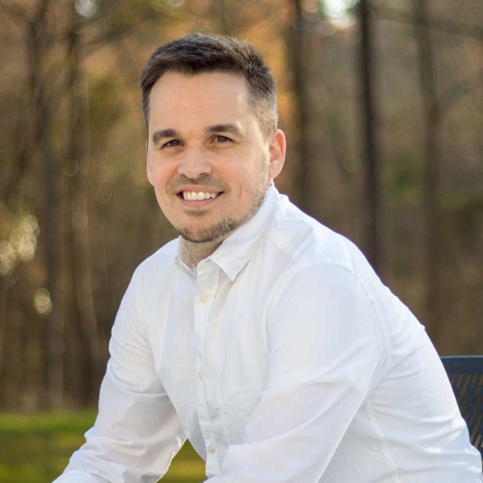A man in a white shirt is sitting on a bench and smiling for the camera.