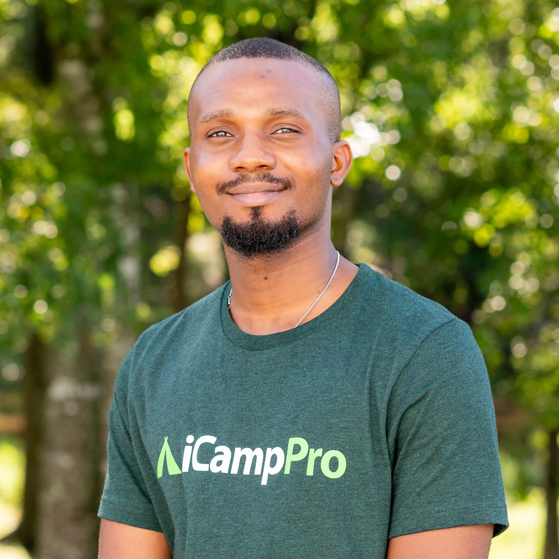 A man with a beard wearing a green t-shirt is standing in front of trees.
