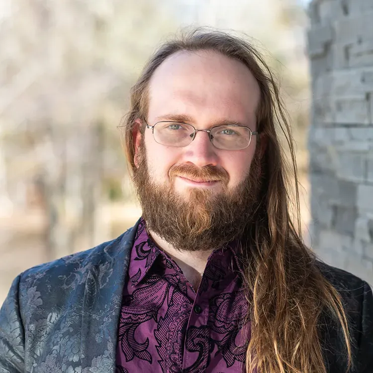 A man with long hair and a beard is wearing glasses and a purple shirt.