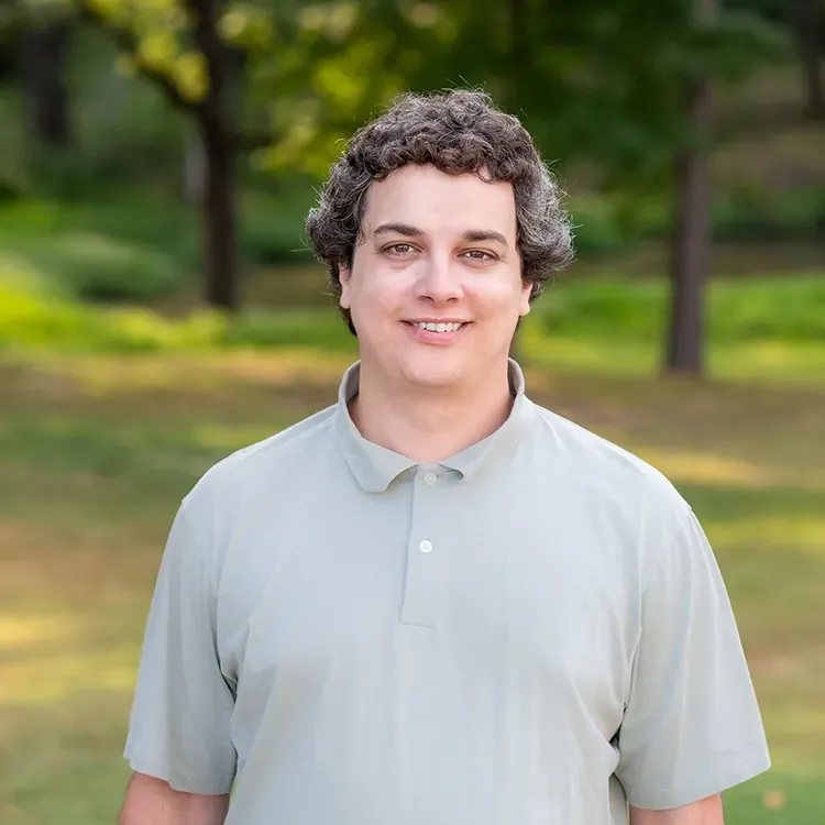 A young man in a grey polo shirt is standing in a park.