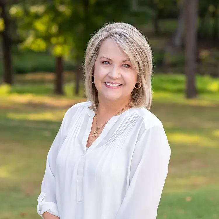 A woman in a white shirt is standing in a park and smiling.
