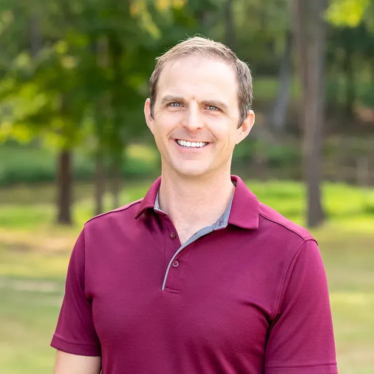 A man in a maroon polo shirt is smiling for the camera in a park.