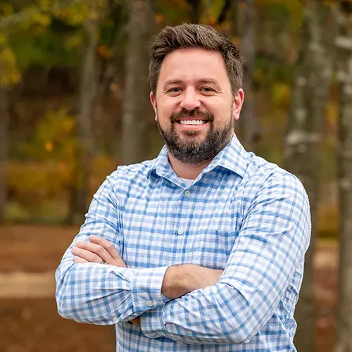 A man with a beard is standing with his arms crossed and smiling.