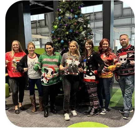 Group of eight people wearing festive sweaters, posing by a decorated Christmas tree in an office setting.