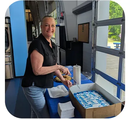 Woman cutting a cake with blue frosting and text. She's smiling, wearing a black shirt and jeans, standing at a blue-covered table.