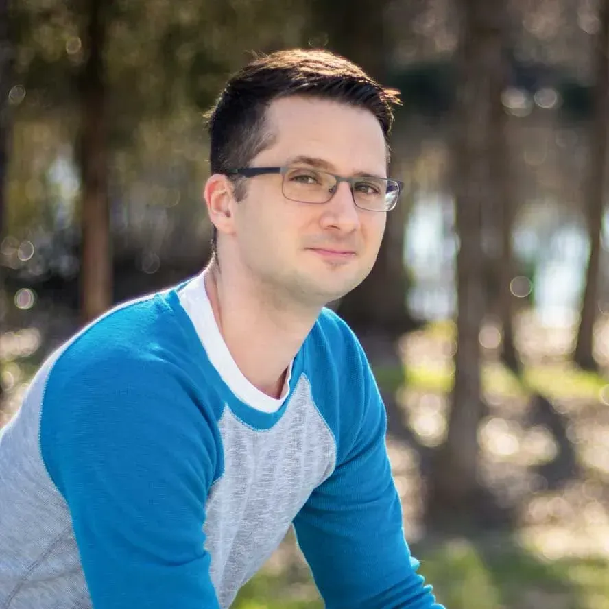 A man wearing glasses and a blue sweater is sitting in the grass.
