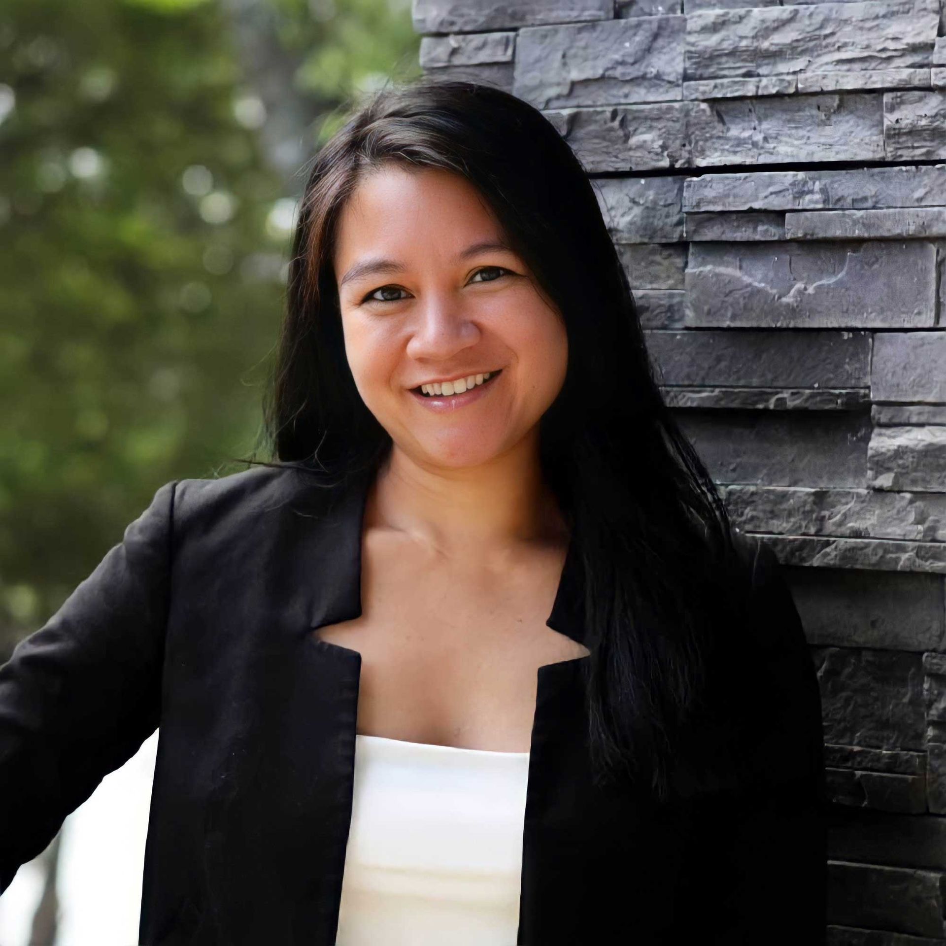 A woman in a black jacket and white top is smiling in front of a stone wall.