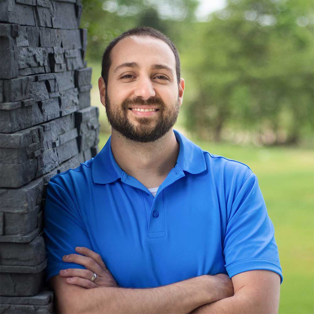 A man with a beard is leaning against a brick wall with his arms crossed.