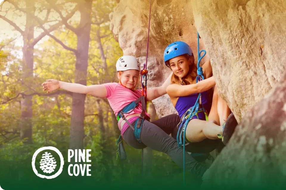 Two girls rock climbing outdoors, smiling, wearing helmets and harnesses.