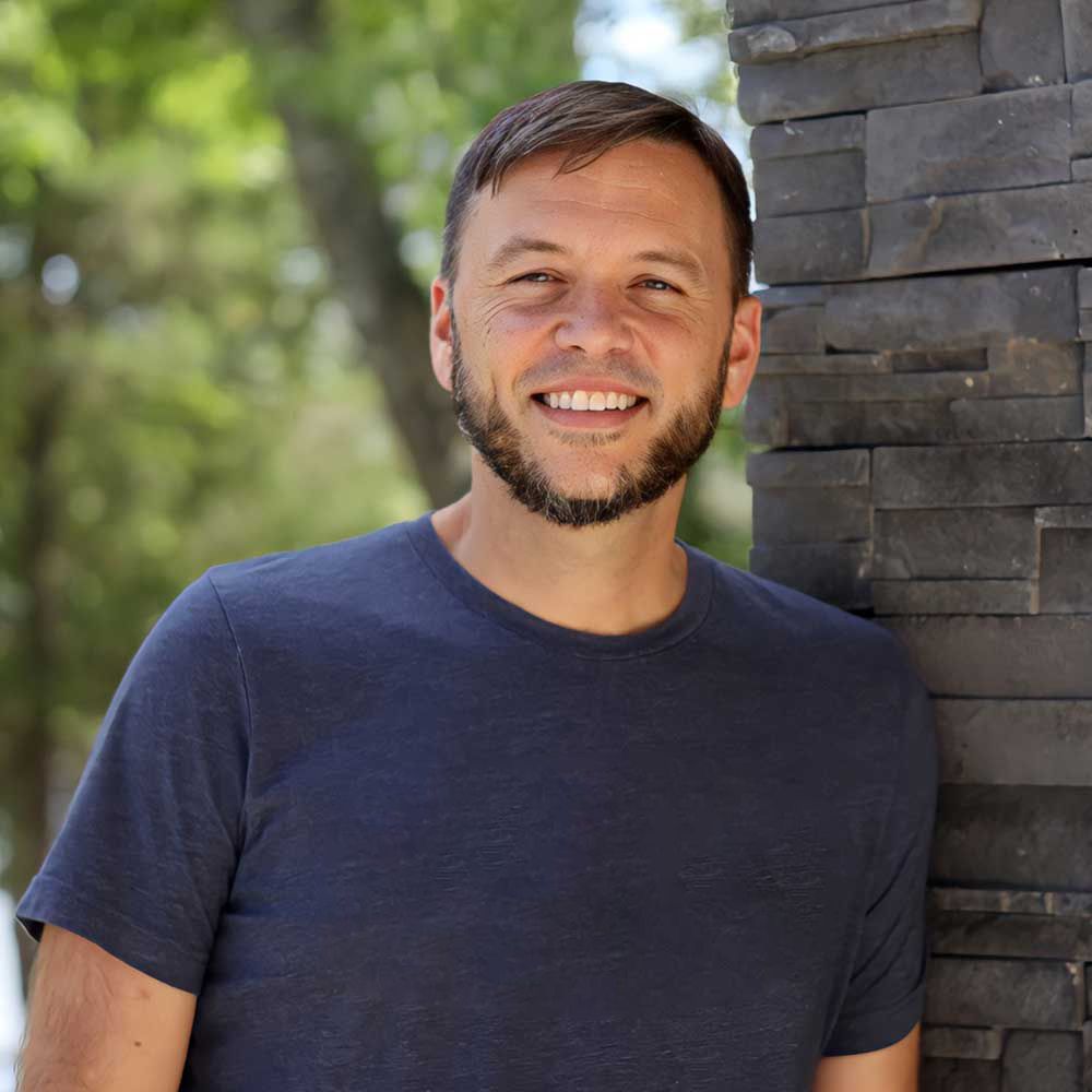 A man with a beard is leaning against a brick wall and smiling.