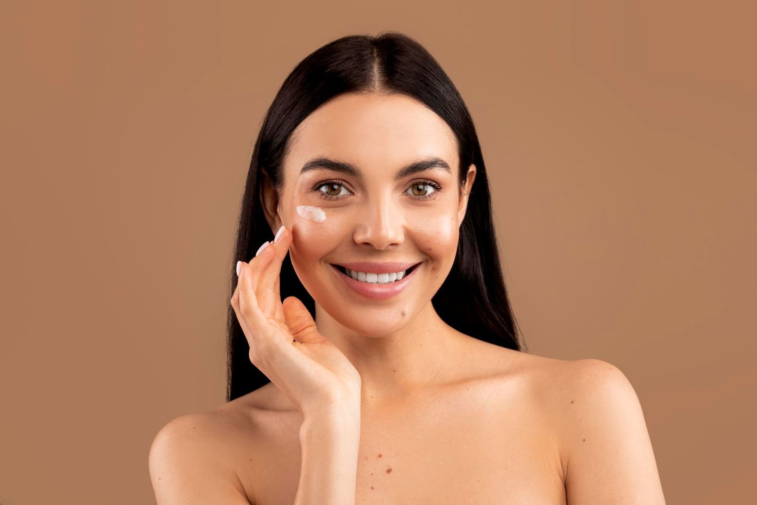 A smiling person applies a small dot of white cream to their cheek against a solid tan background.