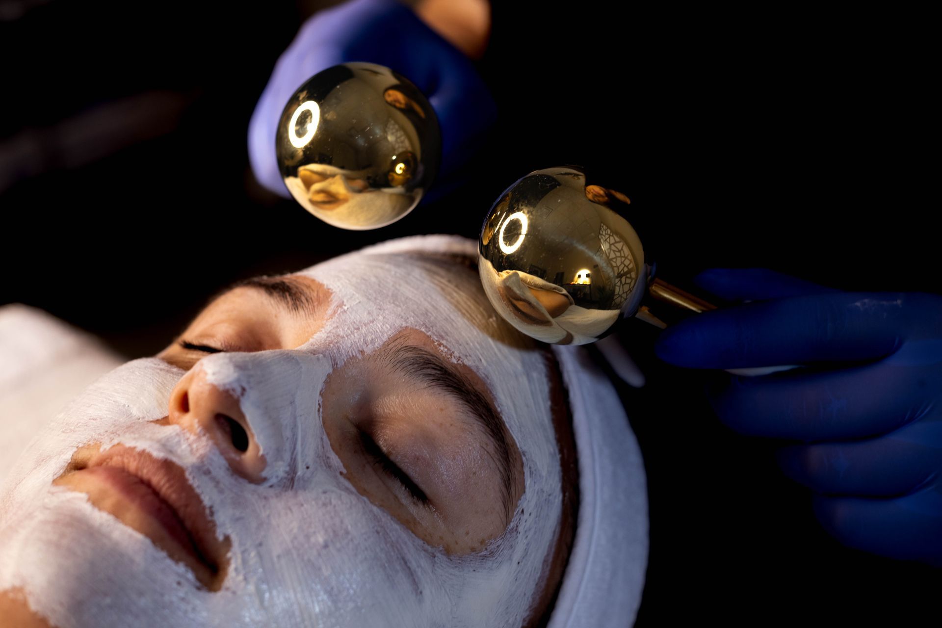 A person with a white facial mask receiving a spa treatment using two gold massage globes held by gloved hands.