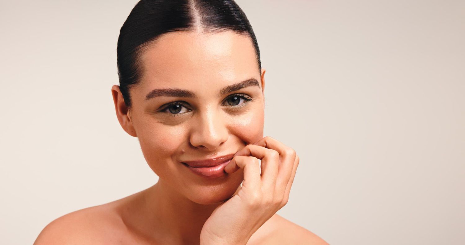 A person with dark hair pulled back smiles softly, resting their chin on their hand against a plain, light-toned background.
