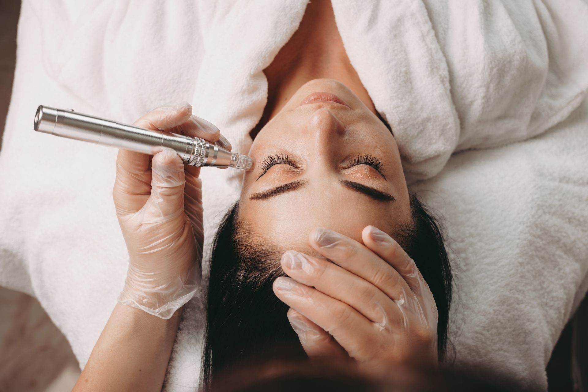 Person receiving facial treatment with a handheld device, lying on a white towel, hands gloved.