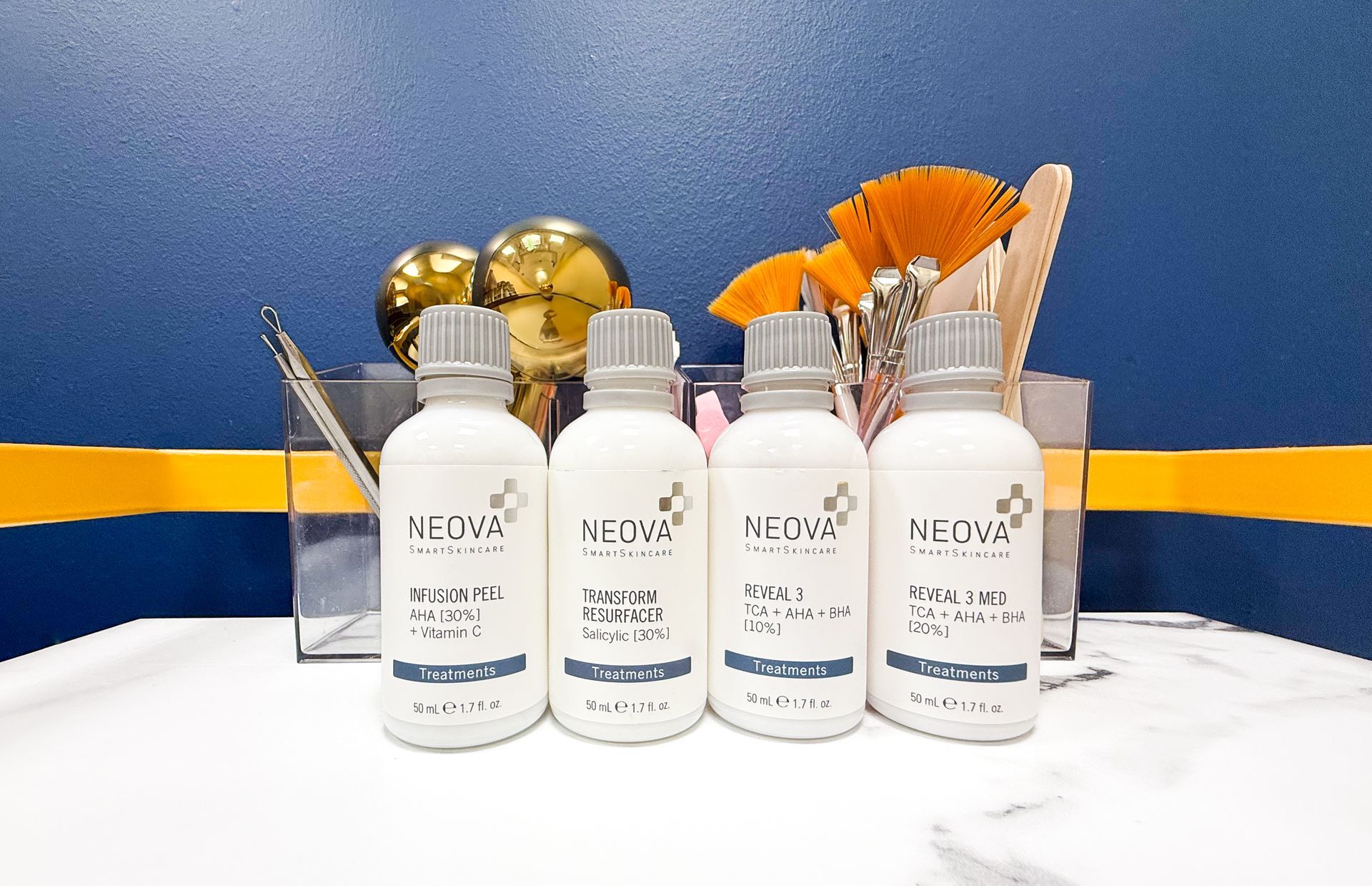 Four white NEOWA skincare bottles aligned on a counter, with brushes and golden decor behind them against a blue wall.