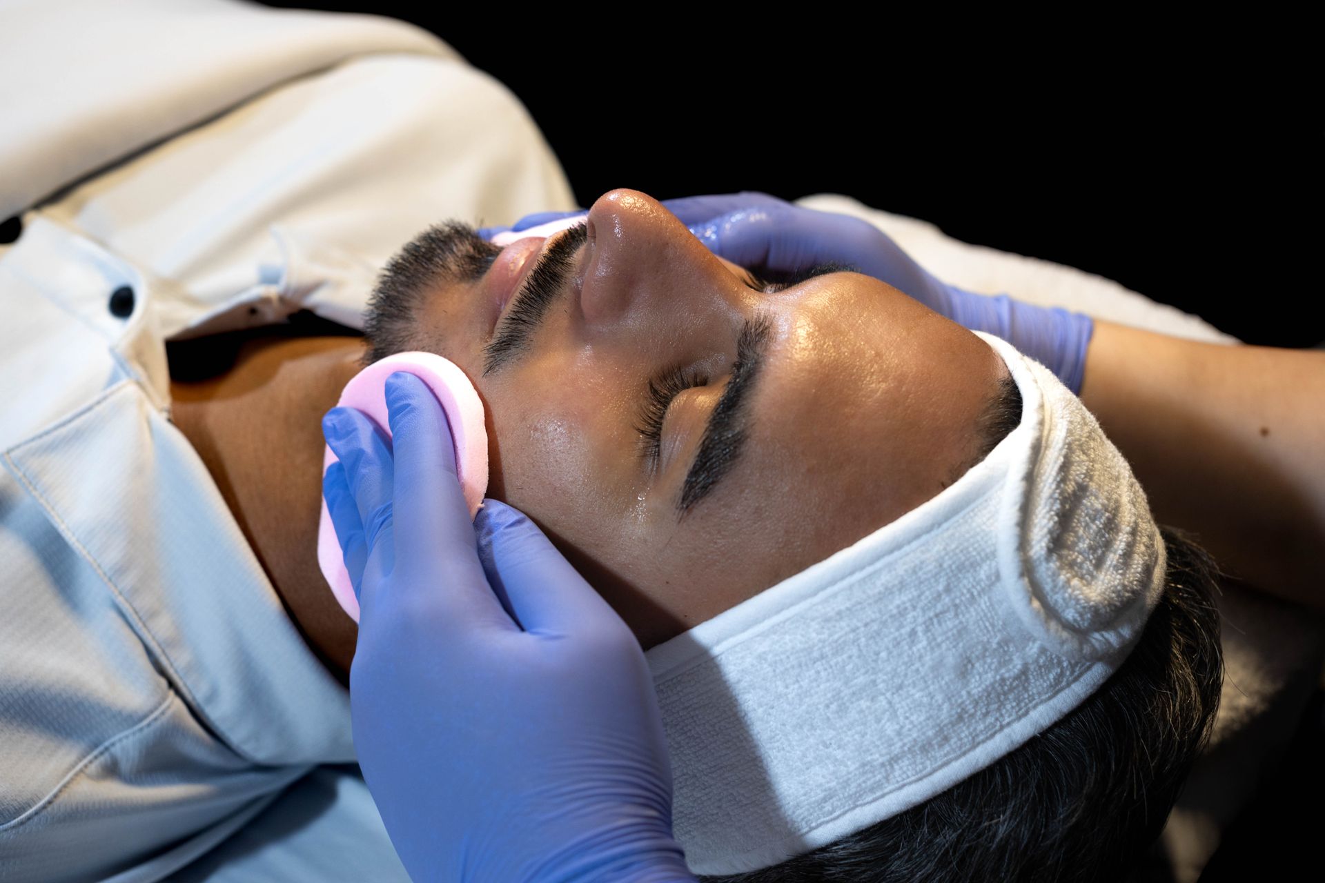 Person receiving facial treatment; hands in blue gloves hold pink sponge and cotton pad on face, wearing a white headband.