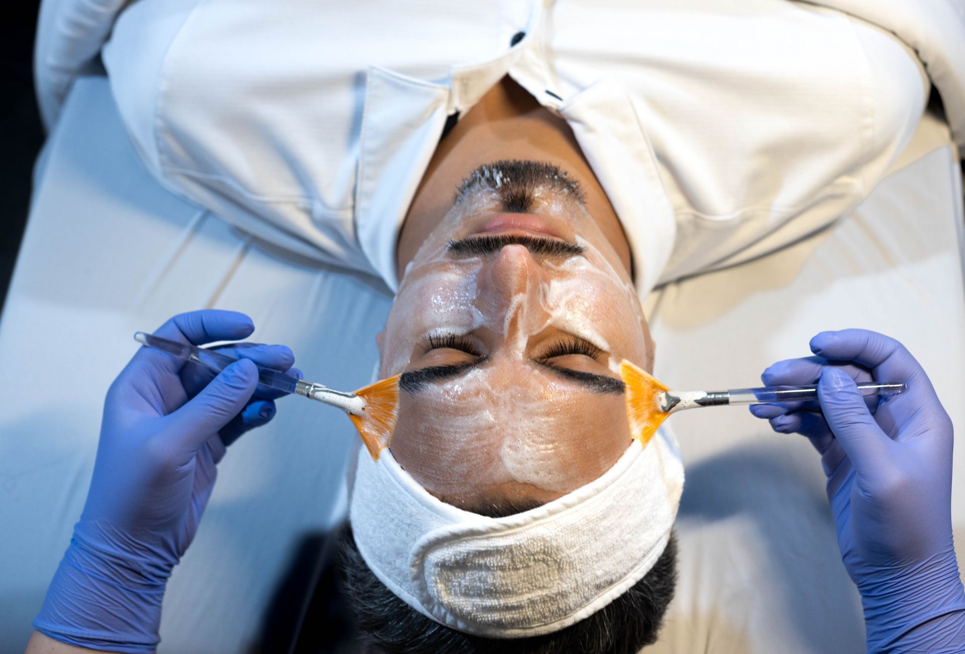 A person receiving facial skin treatment, with a cream mask and tools held by gloved hands.