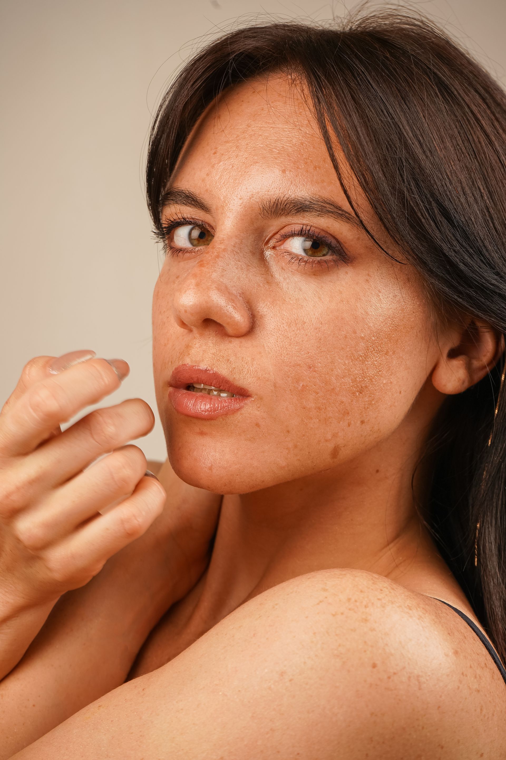 Close-up of a person with dewy, freckled skin and dark hair, looking directly at the camera with a neutral expression.