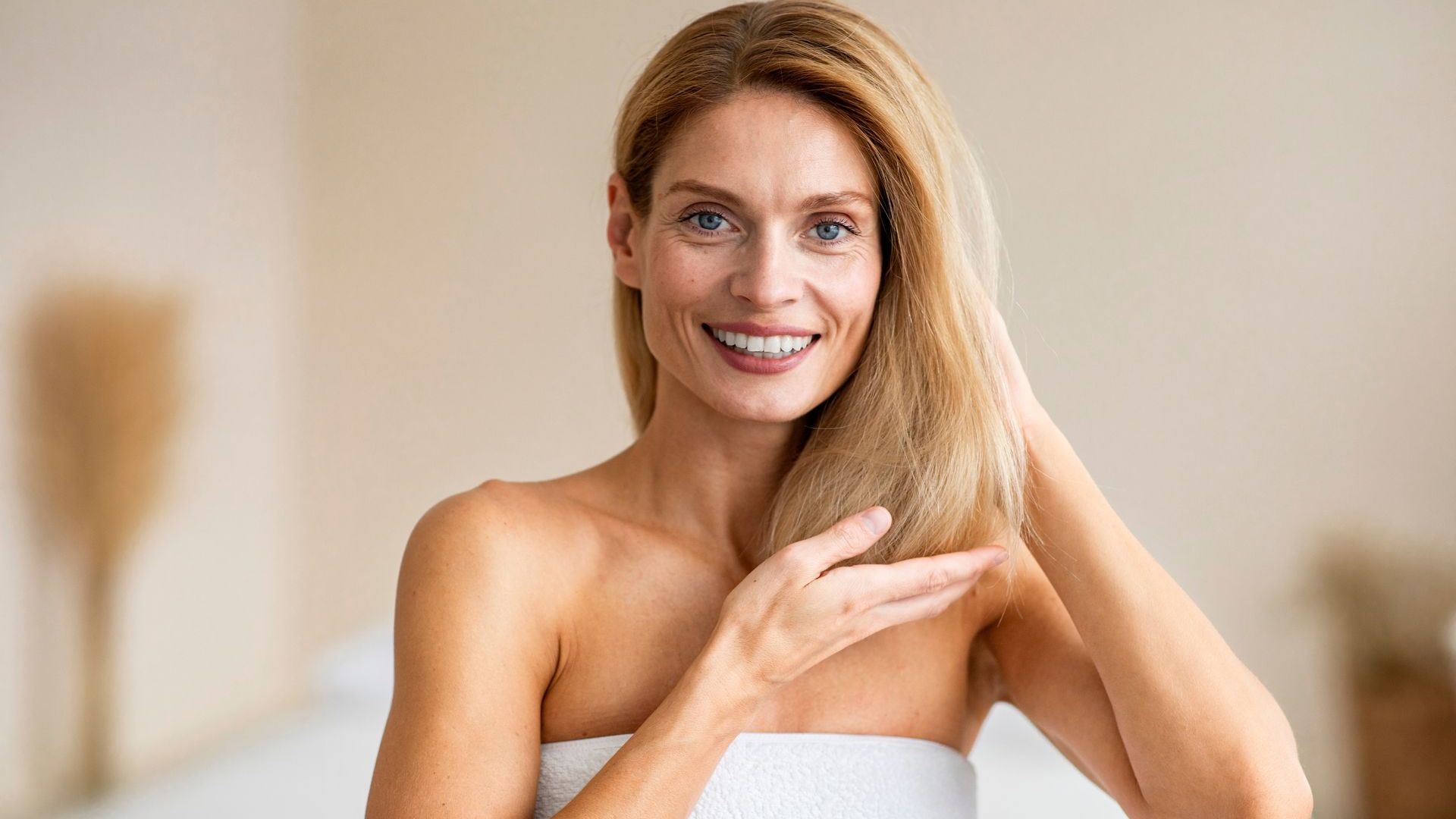 Woman with blonde hair smiles, touching her hair in a light-colored room.