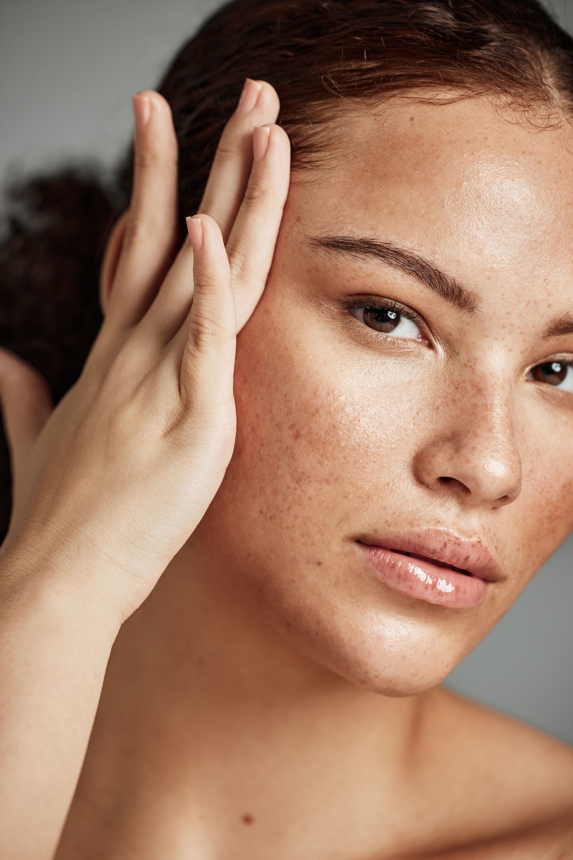 A person with visible skin texture, freckles, and a glossy complexion gently touching the side of their face.