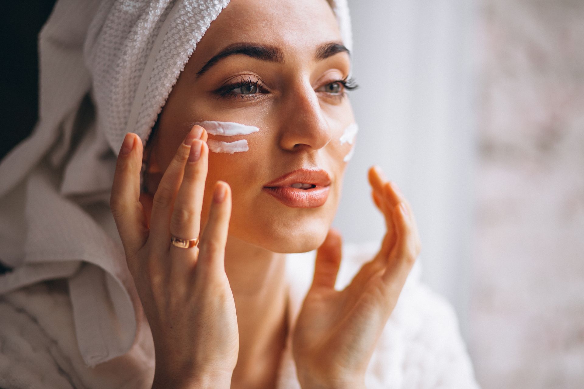 A person wearing a towel wrap applies white cream to their cheeks in a close-up skincare routine shot.
