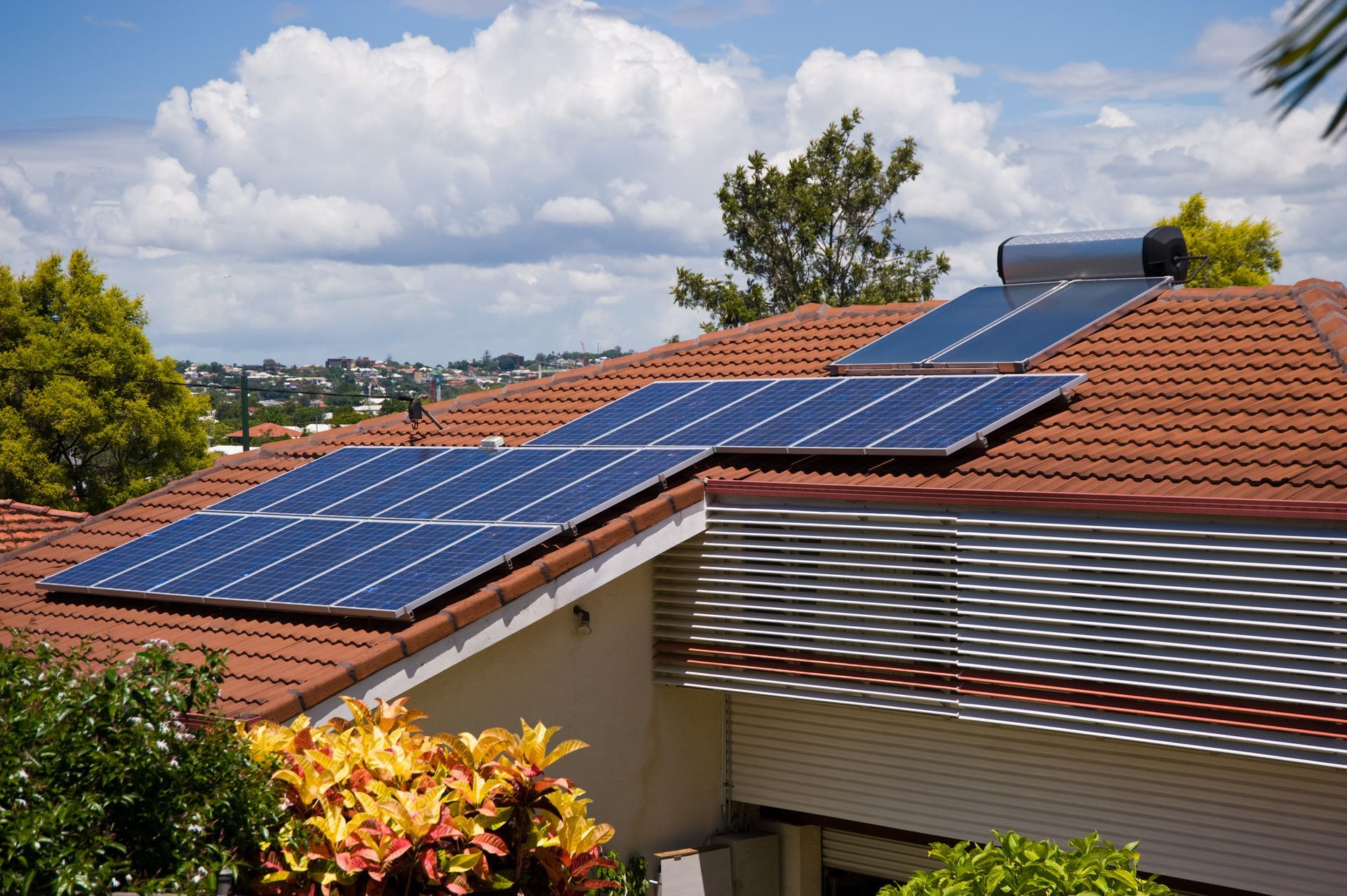 Solar panels on a red-tiled roof with a water heater, plants, and a cityscape in the background.