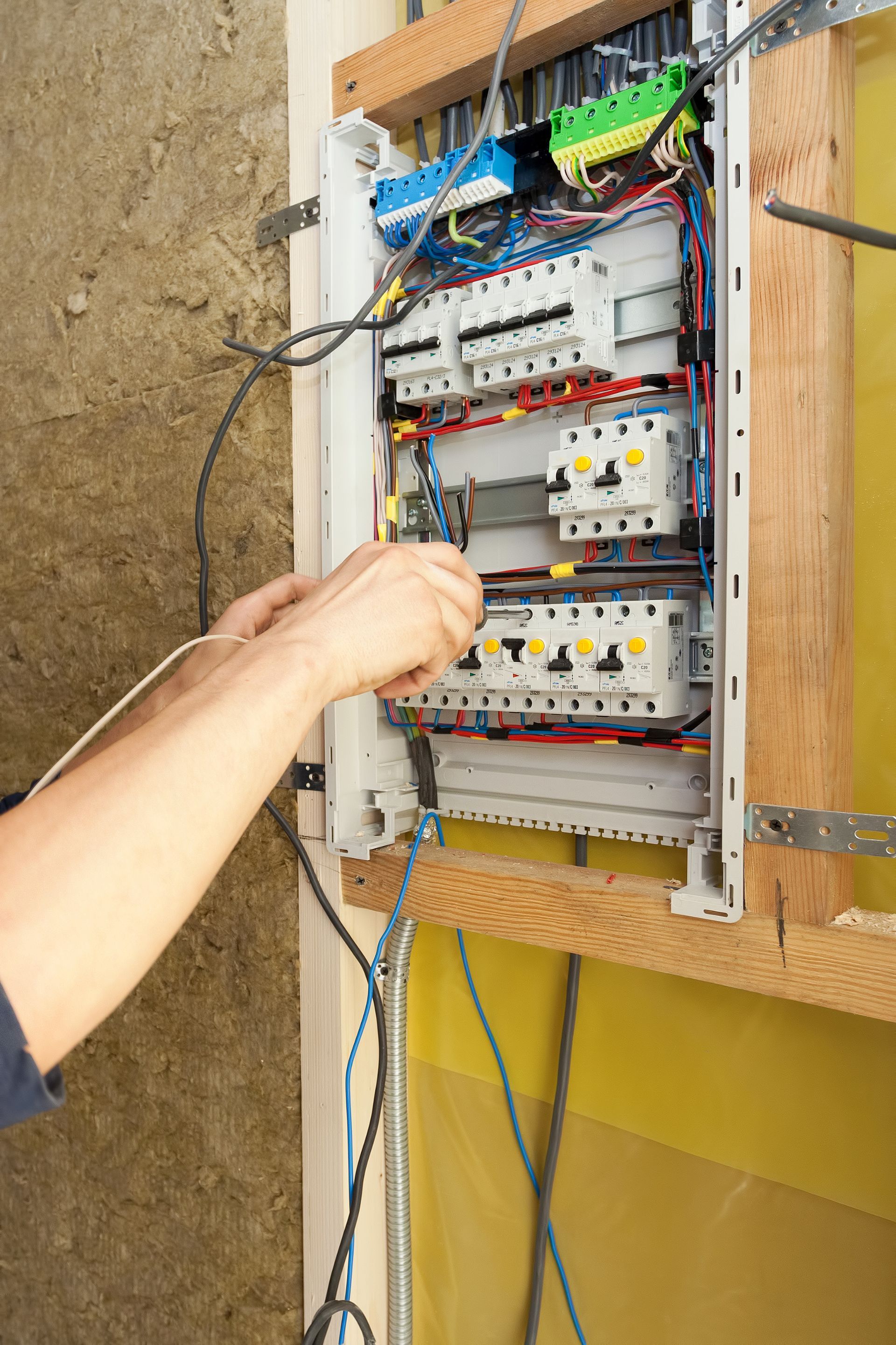 Electrician wiring a breaker box mounted on wood framing near insulation.