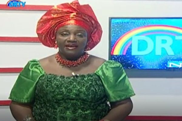 A woman in a green dress and red turban is standing in front of a television.