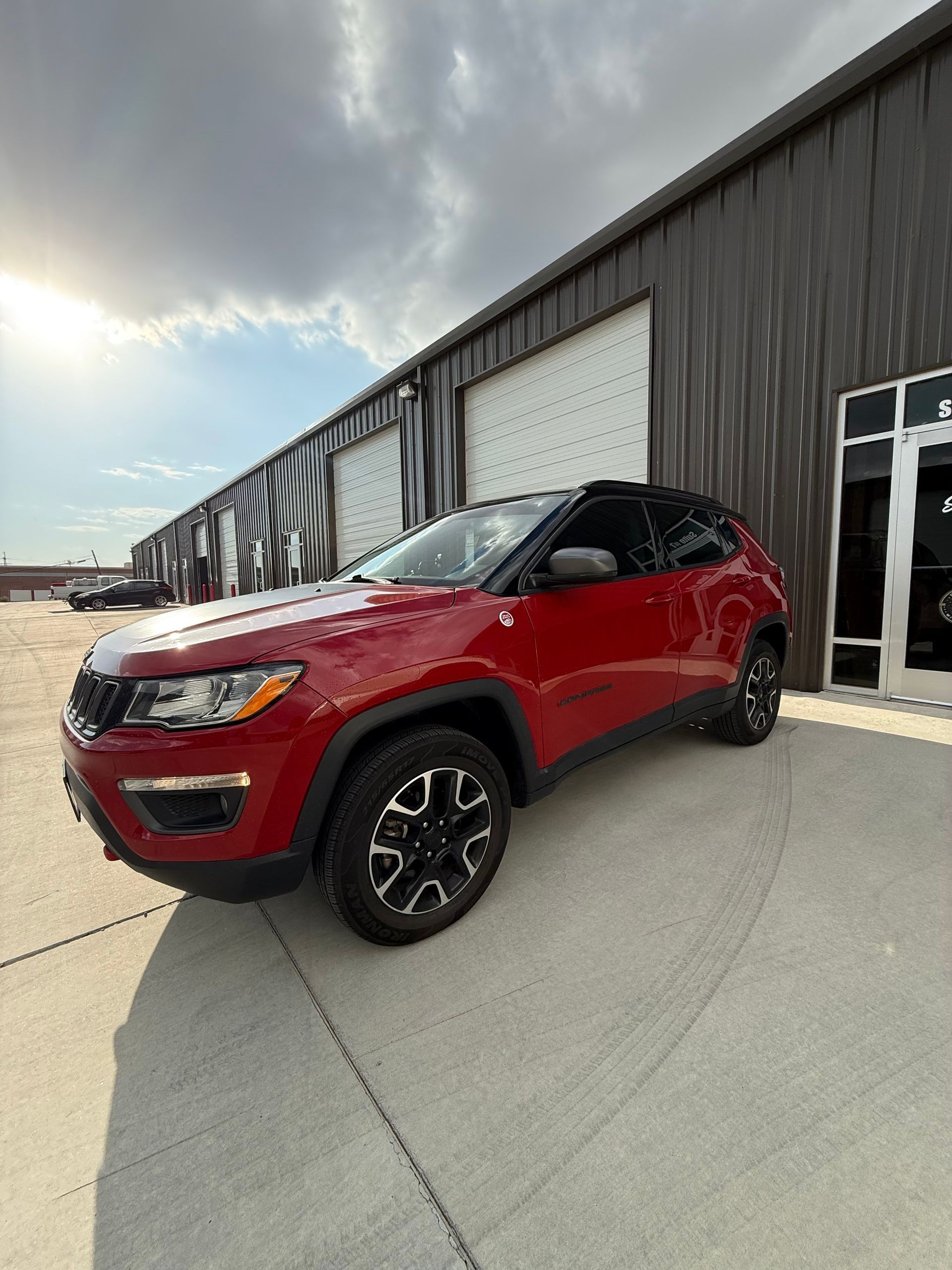 Red Jeep Compass parked in front of a gray industrial building with a cloudy sky.