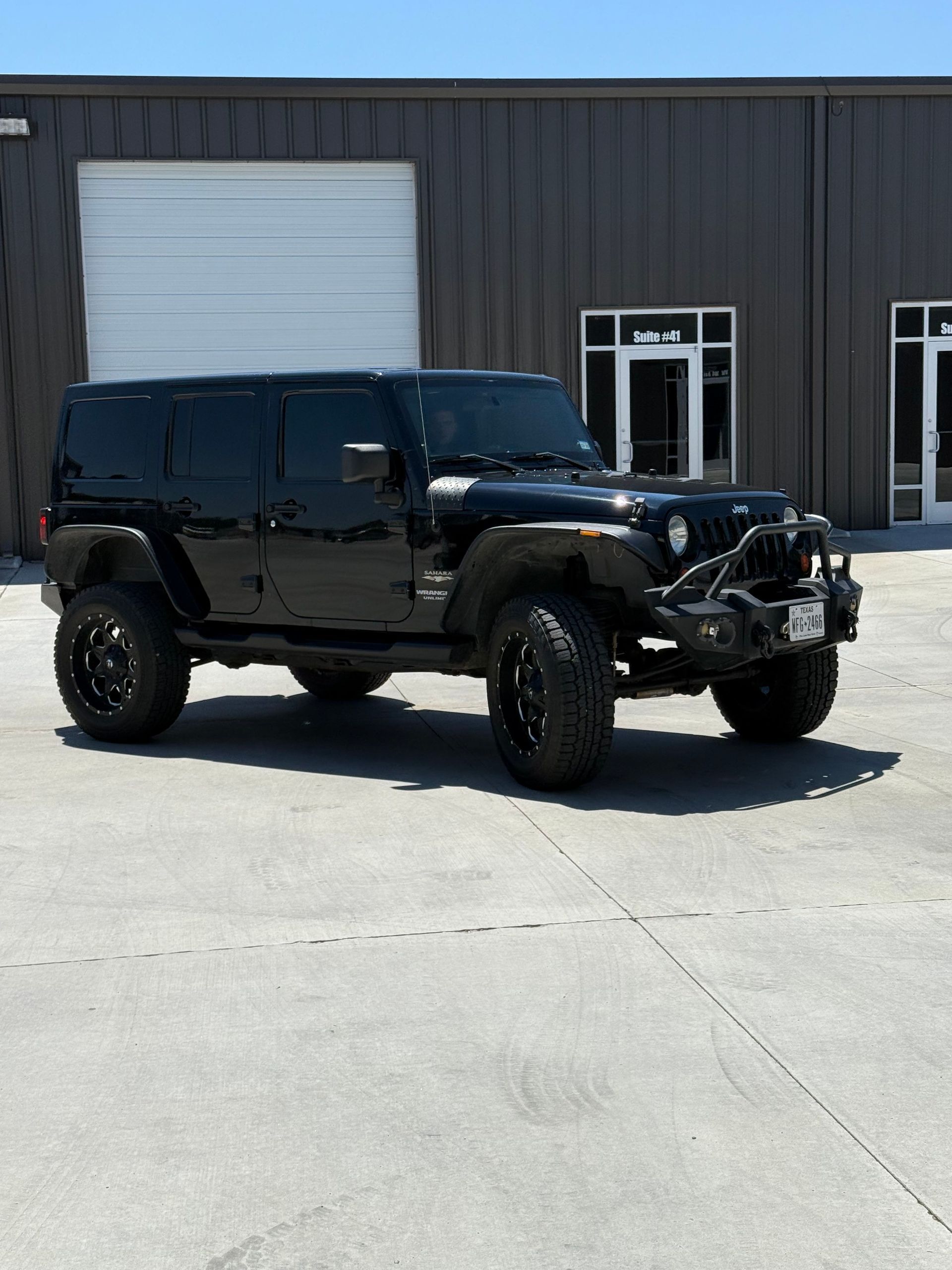 Black Jeep Wrangler parked in front of a gray industrial building on a sunny day.