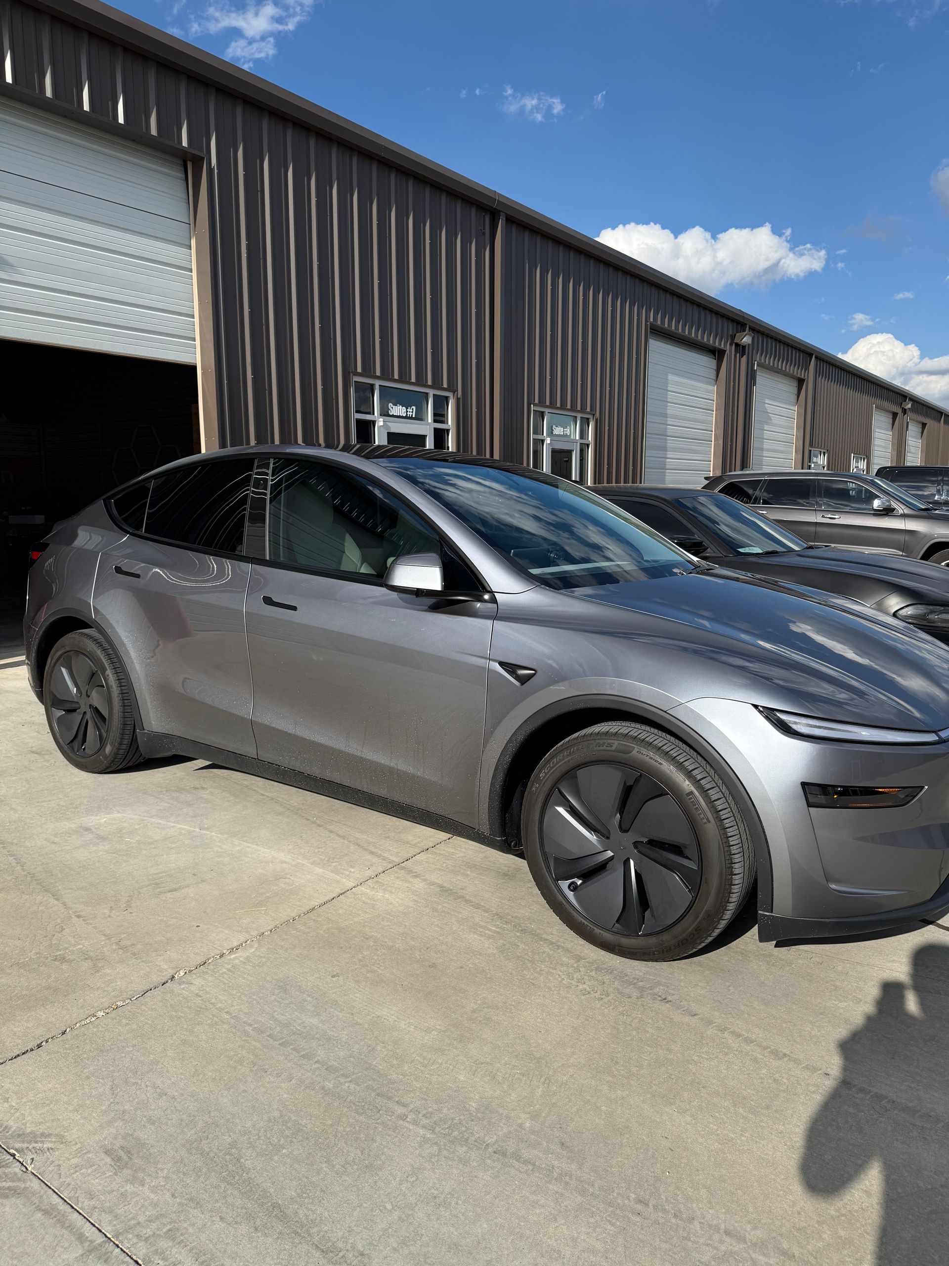 Gray Tesla Model Y parked in front of a metal building with garage doors, sunny day.